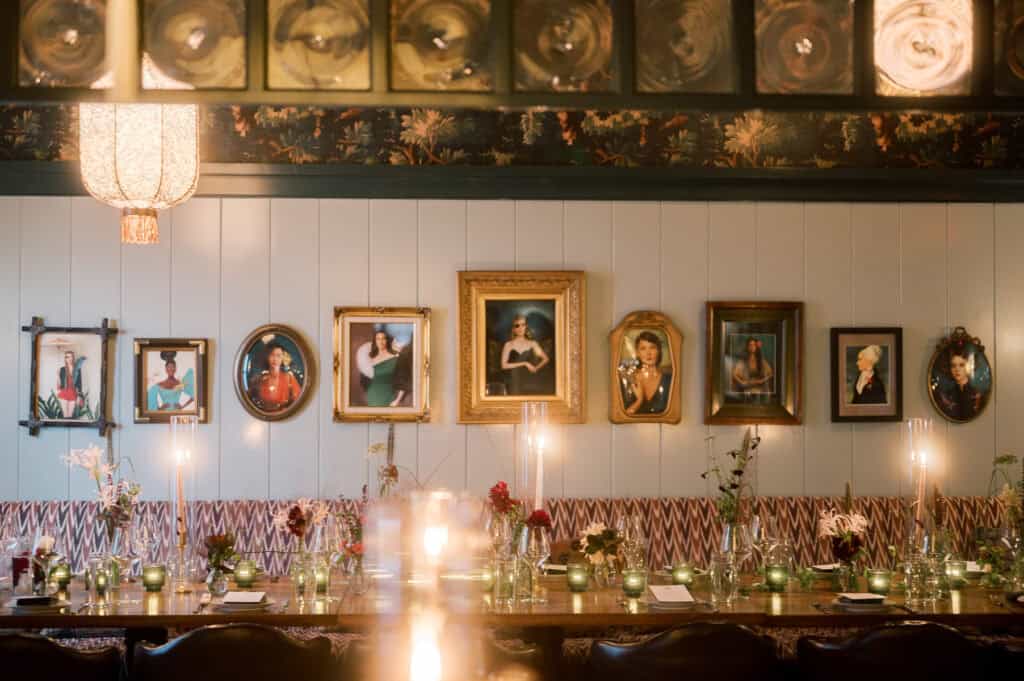 A long dining table at Sister Ship Restaurant in Nantucket is set with candles, flowers, and glassware; framed portraits hang on the wall behind it in a warmly lit room at a Nantucket wedding photographed by Kaity Farrell.