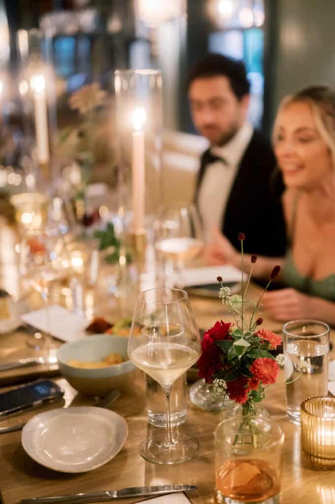 Elegant dinner table with flowers, candles, and glassware, with a man in a suit and a woman in a dress seated and smiling in the softly lit background, captured by Nantucket wedding photographer Kaity Farrell.
