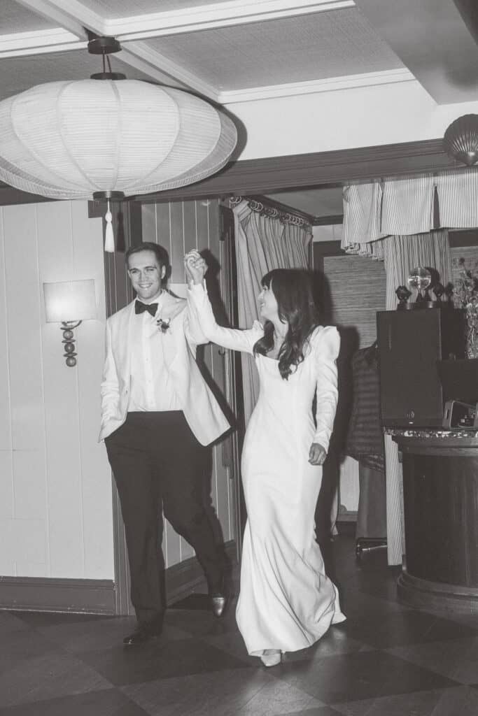 A couple, dressed in formal wedding attire, walks hand in hand into a room, smiling. This black and white image captures a joyful moment from a Nantucket wedding photographed by Kaity Farrell.