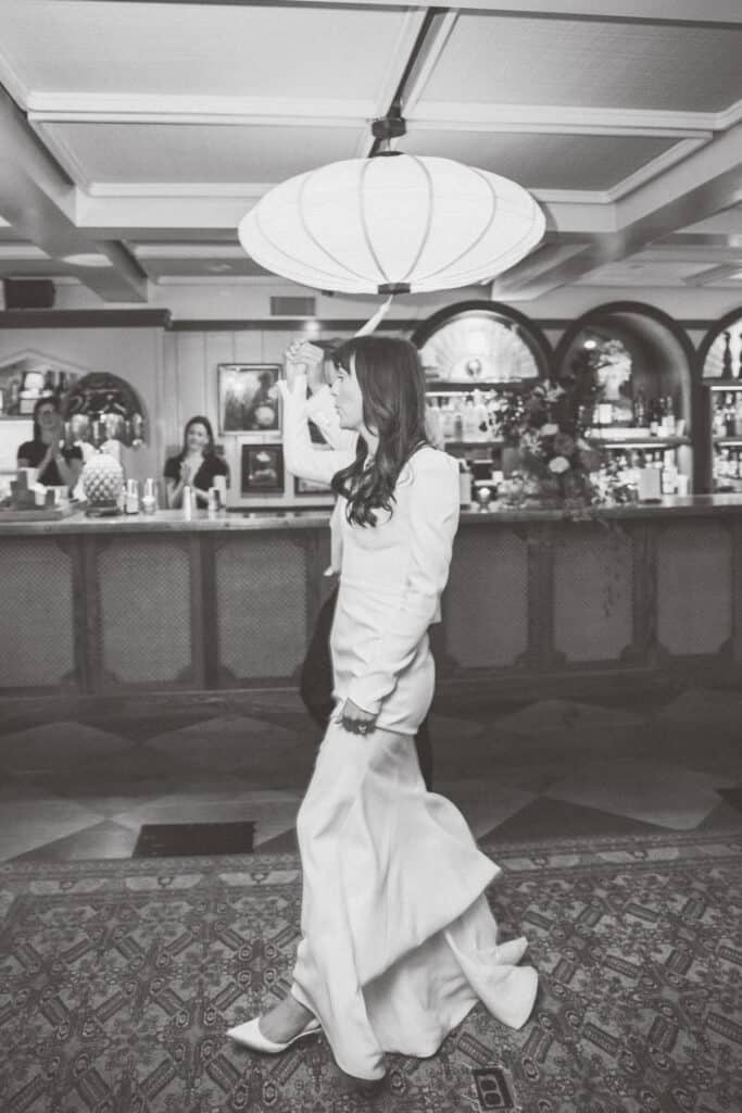 A woman in a long, flowing dress walks indoors, holding a parasol above her head; people and a bar are visible in the background, beautifully captured by Nantucket wedding photographer Kaity Farrell.