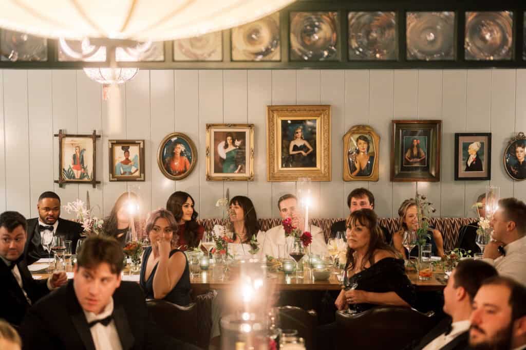 A group of people dressed in formal attire sit at a long dining table in a warmly lit room with framed portraits on the wall behind them, beautifully captured by Nantucket wedding photographer Kaity Farrell.