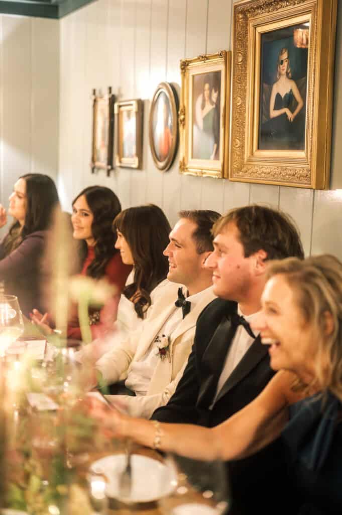 A group of people dressed formally sit at a dinner table, smiling and laughing, with framed portraits hanging on the wall behind them at a Nantucket wedding photographed by Kaity Farrell.