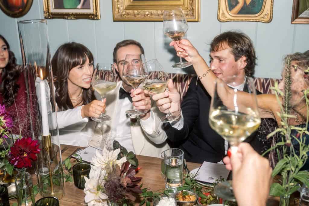 A Nantucket bride and groom and wedding guests dressed formally sit at a decorated table, raising wine glasses in a toast at a Nantucket wedding photographed by Kaity Farrell.