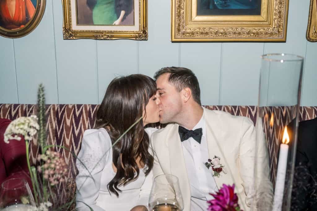 A bride and grrom dressed in formal attire kisses while seated at a table in a decorated room with framed artwork, beautifully captured by Nantucket Wedding Photographer Kaity Farrell.