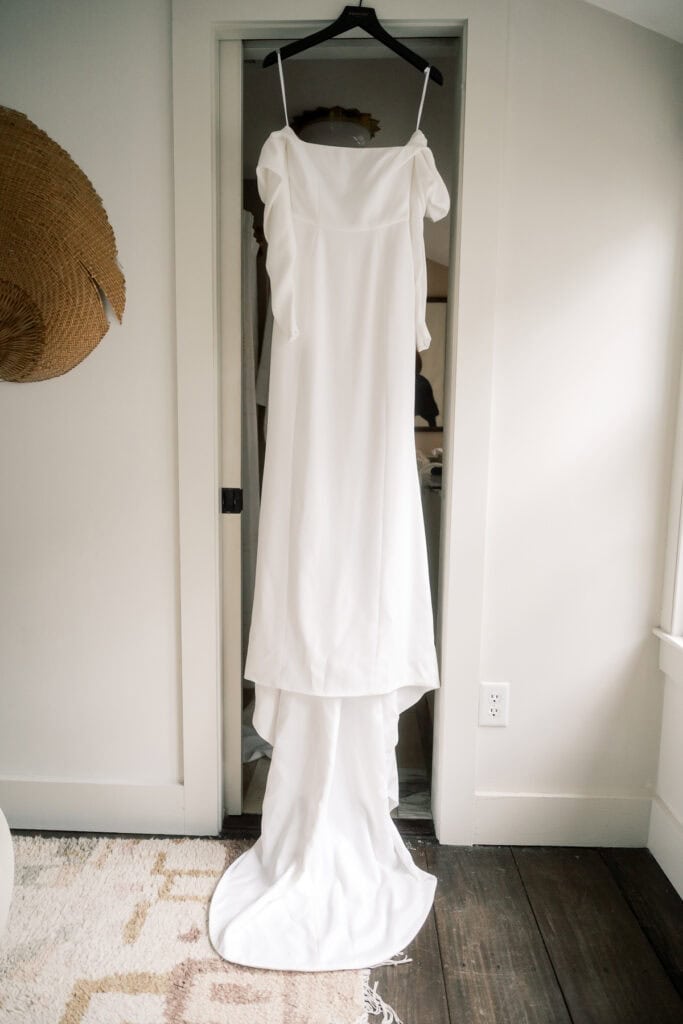 A long, white sleeveless wedding dress with thin straps by Halfpenny London is hanging on a hanger in a doorway inside a bright room, ready for its moment—captured beautifully by Nantucket Wedding Photographer Kaity Farrell.