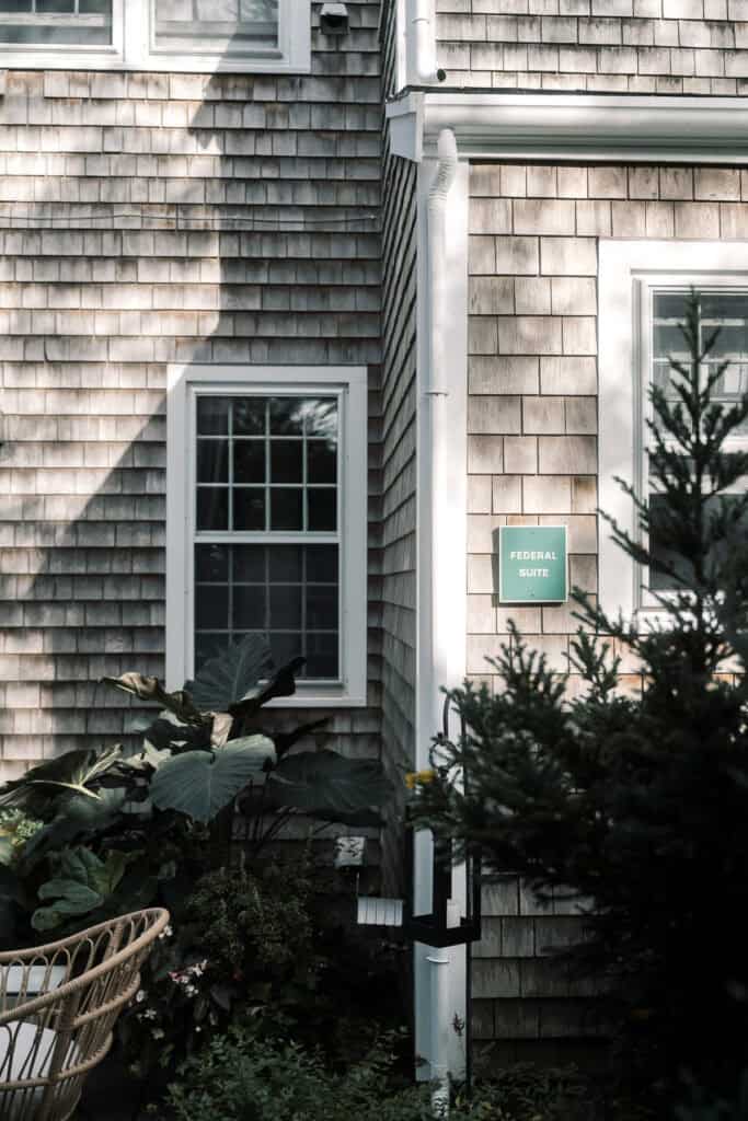 A wooden shingle house exterior with two white-trimmed windows, leafy plants, a wicker chair, and a green "Federal Suite" sign next to a white downspout—captured by Nantucket Wedding Photographer Kaity Farrell.