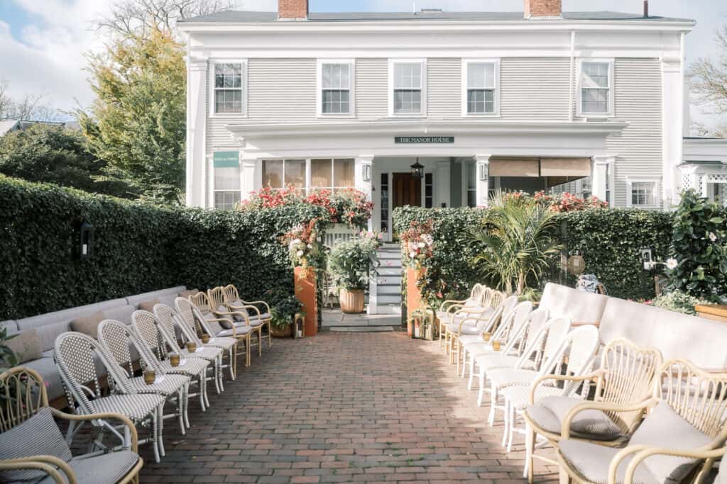 Outdoor courtyard at Faraway Nantucket hotel with rows of wicker chairs facing each other, a brick pathway, lush greenery, and a two-story white house in the background—perfectly captured by Nantucket Wedding Photographer Kaity Farrell.