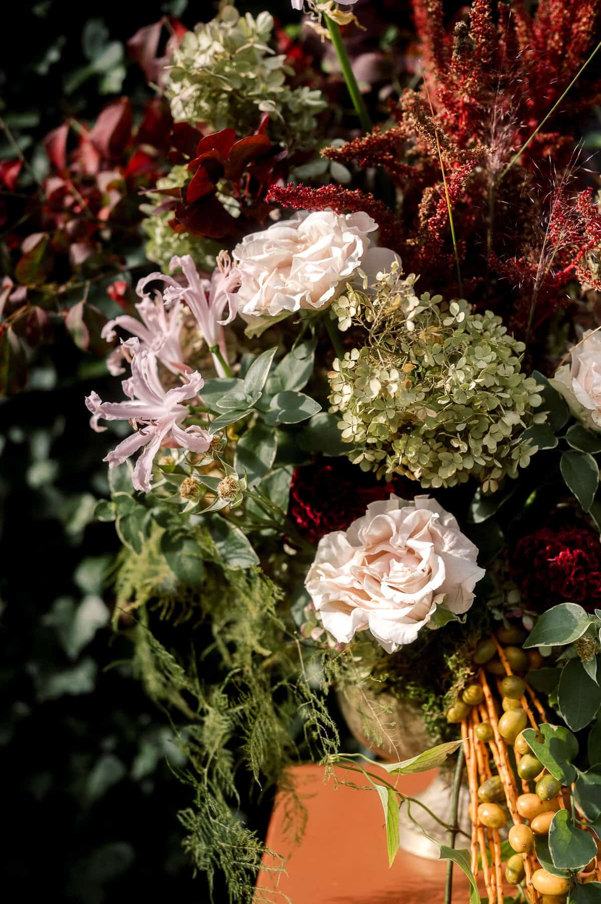 A floral arrangement inspired by a Nantucket wedding photographed by Kaity Farrell, featuring white roses, light pink lilies, green hydrangeas, red flowers, assorted foliage, and clusters of yellow berries.