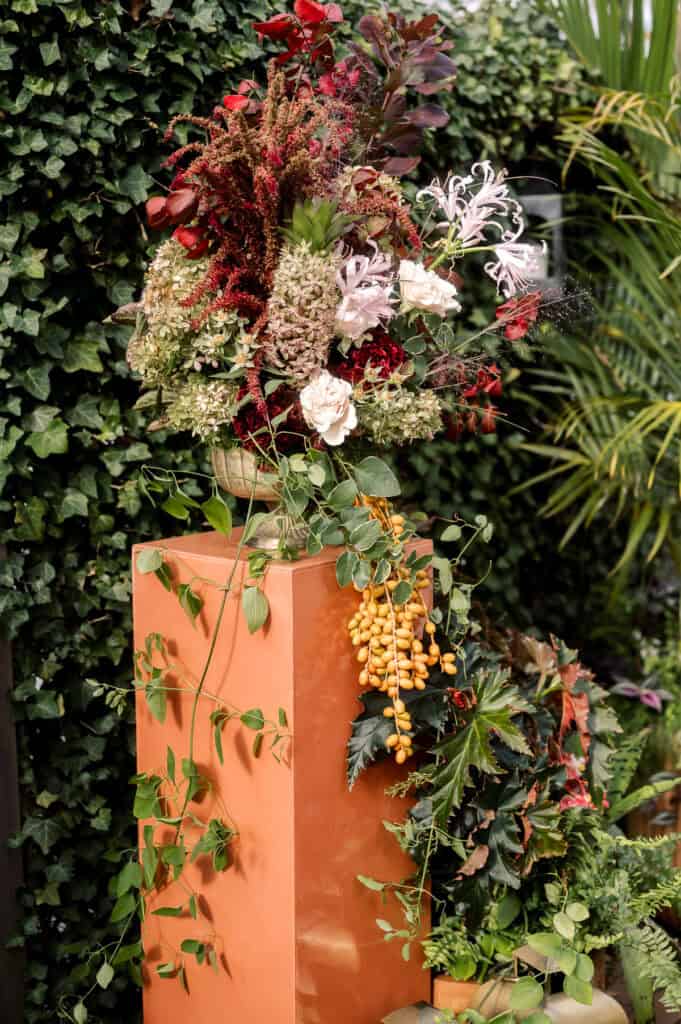 A large floral arrangement with red, white, and pink flowers, leafy greenery, and yellow berries sits on a glossy orange pedestal, surrounded by lush plants—perfect for a Nantucket wedding photographed by Kaity Farrell.