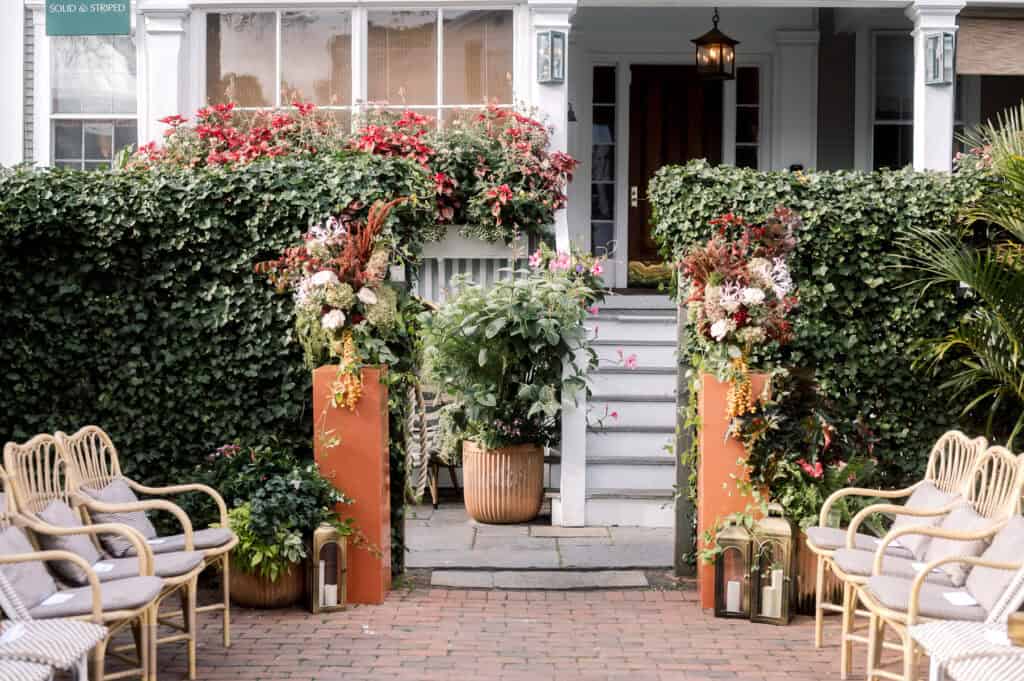 A brick pathway leads to steps up to a house entrance, flanked by tall flower arrangements—an inviting setting for a wedding ceremony—surrounded by green plants and wicker chairs, captured beautifully by Nantucket Wedding Photographer Kaity Farrell.
