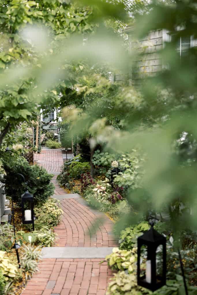 A brick pathway winds through a lush garden with green plants and black lanterns, partially obscured by tree branches in the foreground at a Nantucket wedding at Faraway Nantucket hotel photographed by Kaity Farrell.