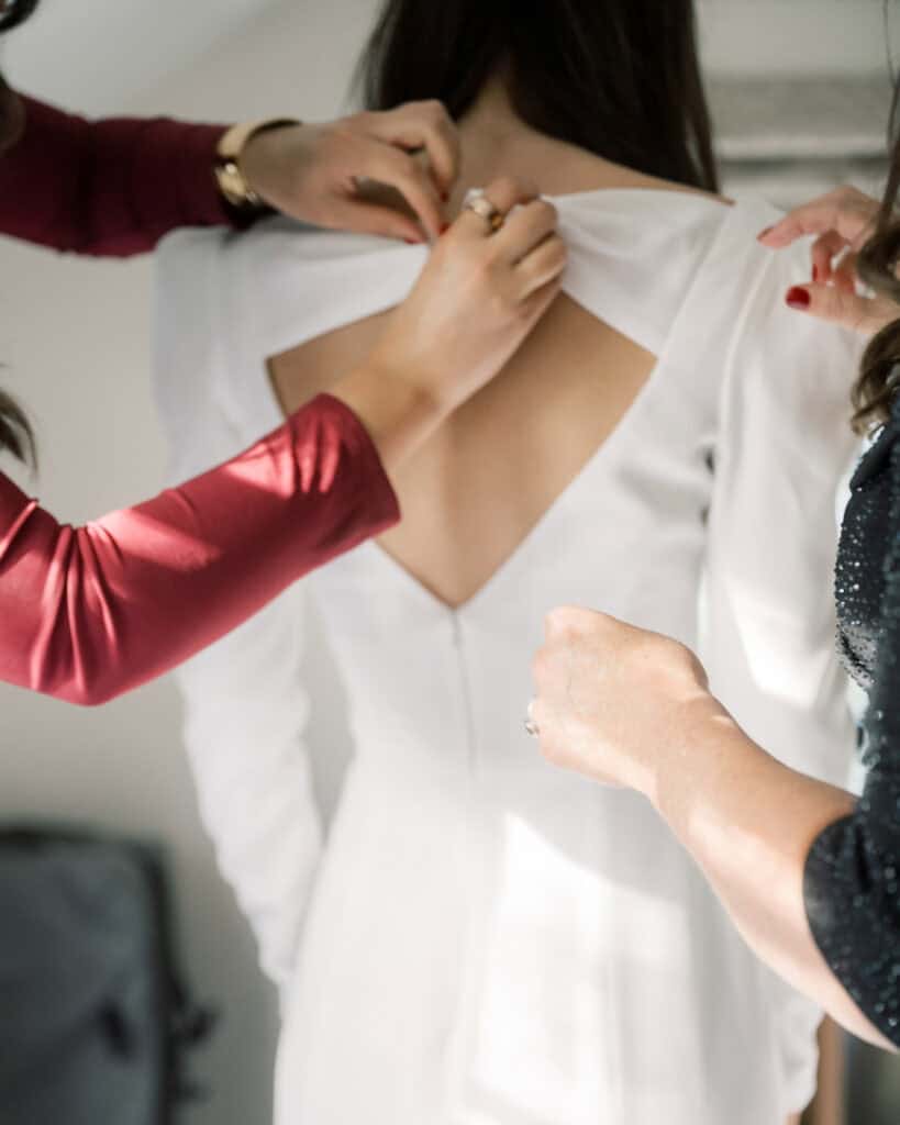 Nantucket Wedding Photographer Kaity Farrell captures two people helping to fasten the back of a bride’s white dress, focusing on her upper back and hands delicately adjusting the fabric.