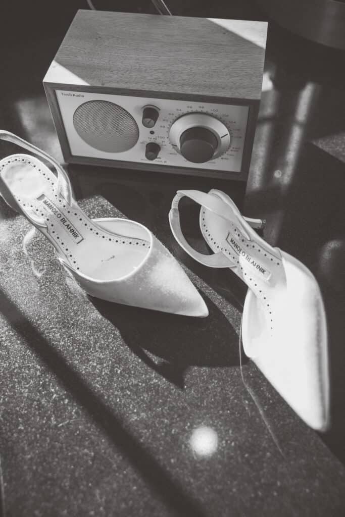 The bride's high-heeled shoes and a vintage-style radio are placed on a dark, glossy surface in sunlight, reminiscent of the timeless elegance captured by Nantucket Wedding Photographer Kaity Farrell.