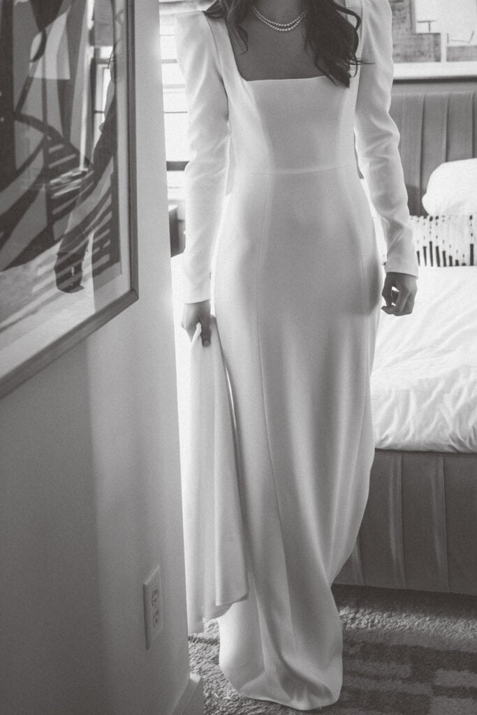 A bride in a long-sleeved, floor-length white dress stands indoors near a bed, holding part of the dress in one hand. Captured in black and white by Nantucket Wedding Photographer Kaity Farrell.