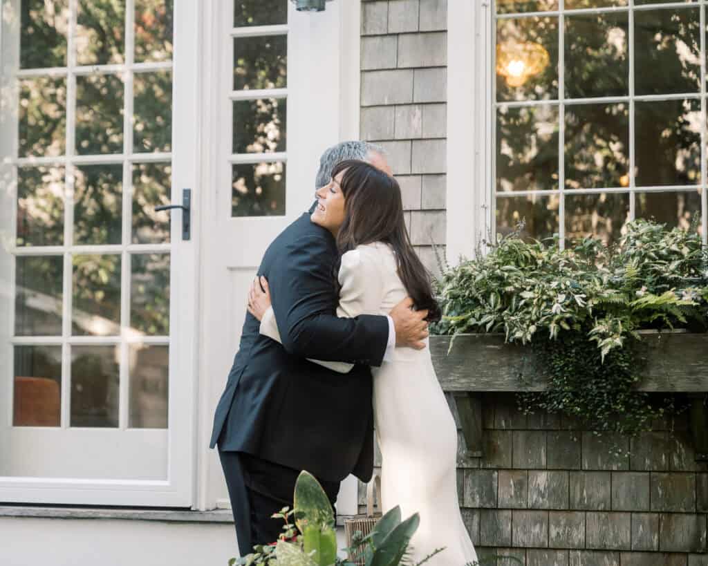 A man in a suit and a woman in a white dress hug each other outside a house with large windows and plants in the foreground.