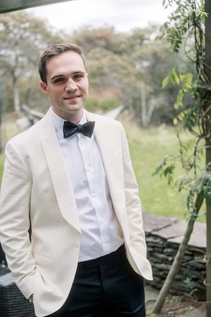 A groom in a white tuxedo jacket, black bow tie, and black pants stands outdoors near greenery, looking at the camera with his hands in his pockets. Captured beautifully by Nantucket wedding photographer Kaity Farrell.