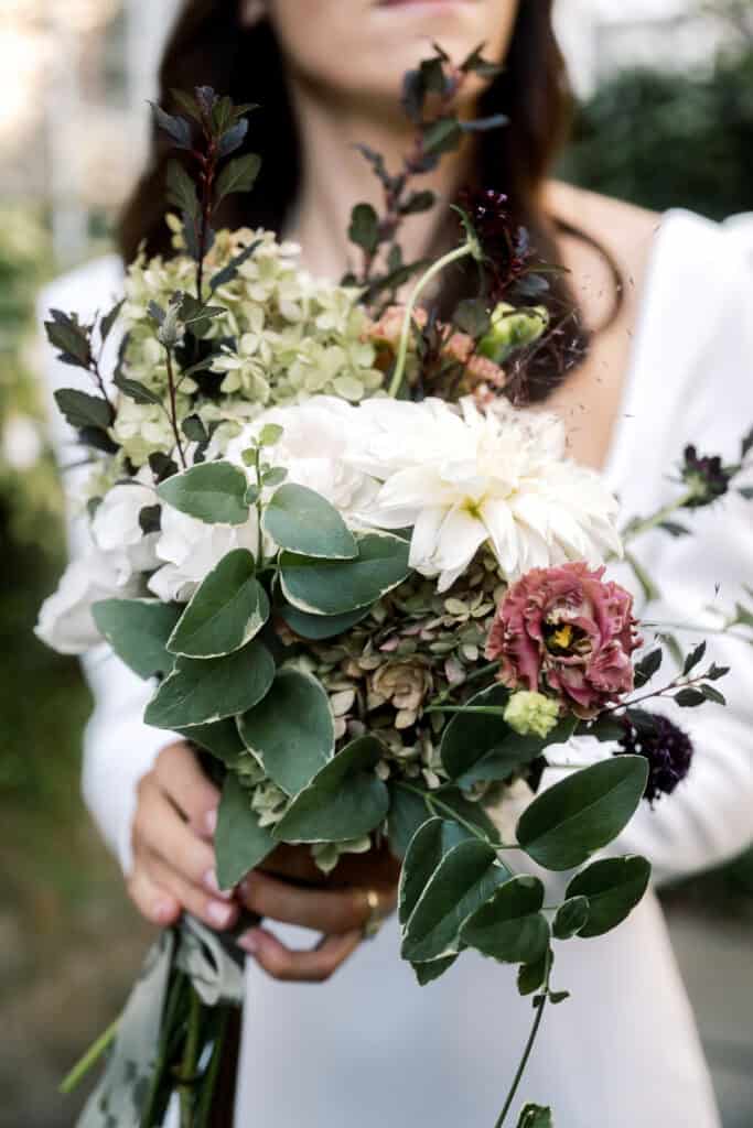 A bride in a white dress holds a bouquet of mixed flowers and greenery, including white, pink, and purple blooms at a Nantucket wedding photographed by Kaity Farrell.
