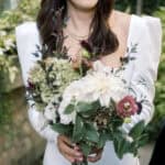A bride in a white dress holds a bouquet of assorted flowers, including white and purple blooms, while smiling slightly outdoors—captured beautifully by Nantucket wedding photographer Kaity Farrell.