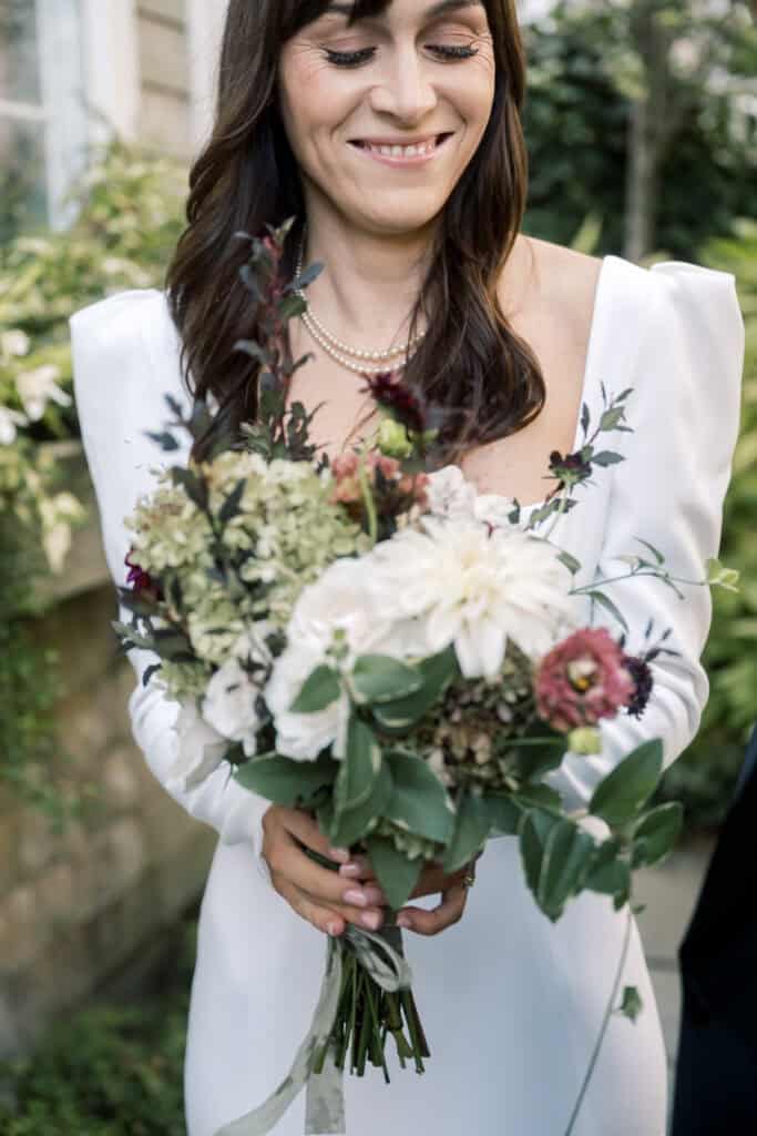 A bride in a white dress holds a bouquet of assorted flowers, including white and purple blooms, while smiling slightly outdoors—captured beautifully by Nantucket wedding photographer Kaity Farrell.
