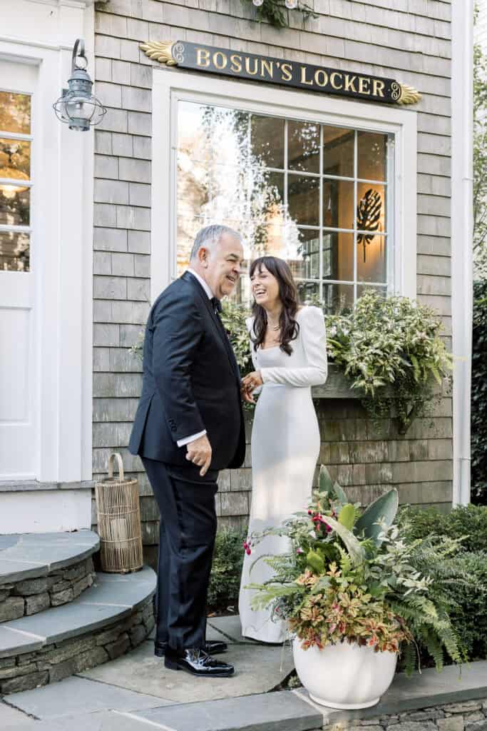 Father of the bride in a suit and the bride in a long white dress stand smiling together outside a house with a sign reading “BOSUN’S LOCKER” above the window at a Nantucket wedding photographed by Kaity Farrell