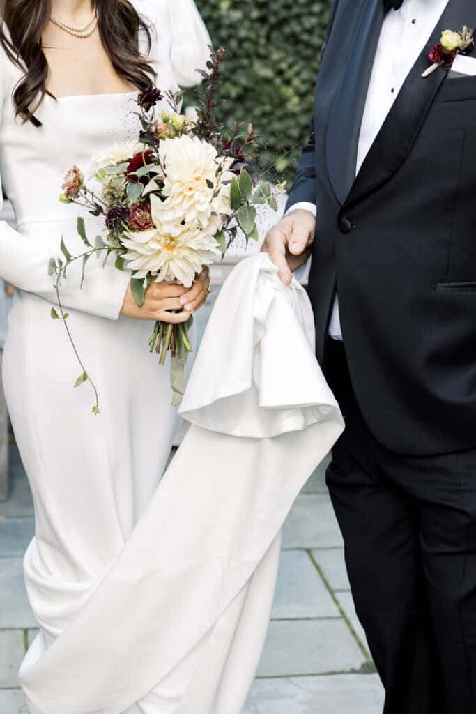 A bride in a white dress holds a bouquet of flowers while standing next to her father in a black tuxedo holding part of her dress, captured beautifully by Nantucket Wedding Photographer Kaity Farrell.