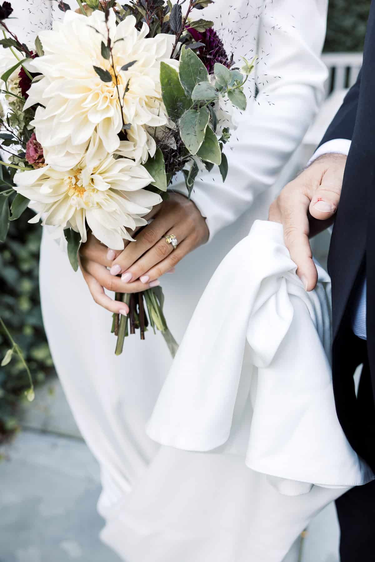 A Nantucket bride in a white dress holds a bouquet of flowers, showing a ring, as her father in a suit holds part of the dress—captured beautifully by Nantucket Wedding Photographer Kaity Farrell.