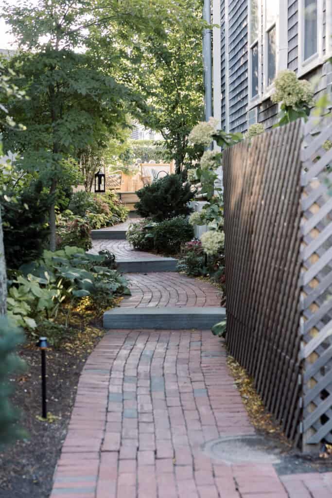 A curved brick pathway leads through a garden beside a building with wooden siding and a lattice fence, surrounded by plants and trees.