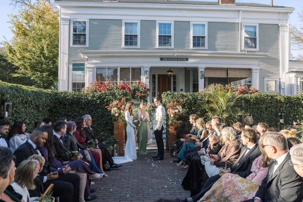 A wedding ceremony takes place outdoors in front of a white house, with guests seated on either side of the aisle watching the couple and officiant, beautifully captured by Nantucket Wedding Photographer Kaity Farrell.