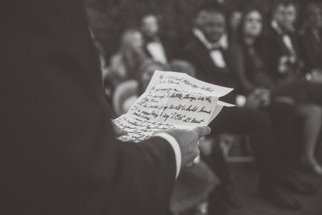 A person in a suit holds handwritten notes while standing in front of a seated guests dressed in formal attire at a Nantucket wedding, captured candidly by Nantucket Wedding Photographer Kaity Farrell.