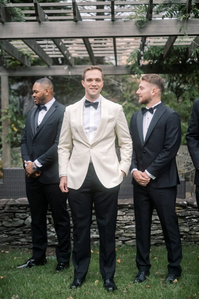 Three men in tuxedos stand beneath a wooden pergola in a garden; the groom in the center wears a white jacket, while the others wear black. Captured by Nantucket wedding photographer Kaity Farrell.