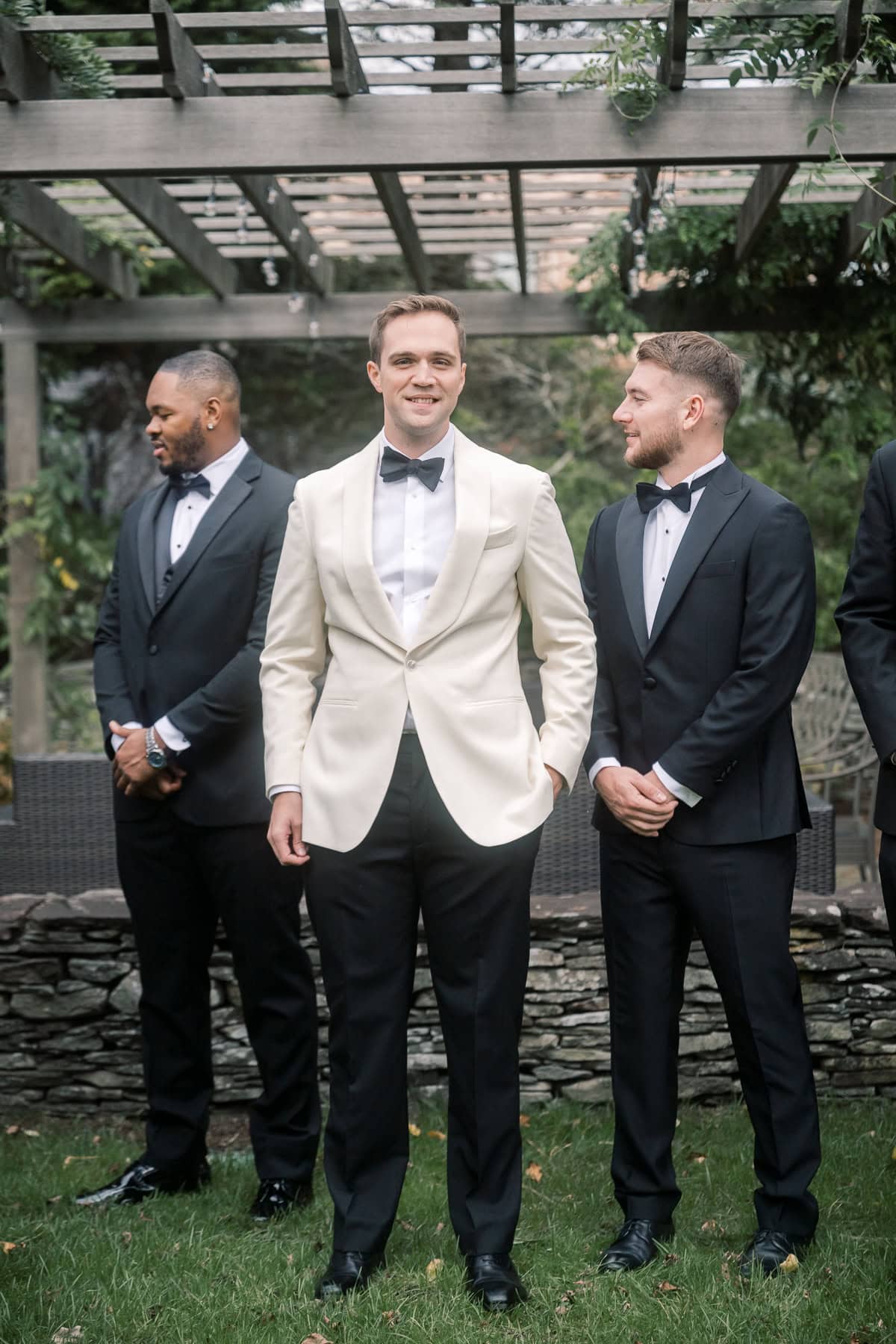 Three men in tuxedos stand beneath a wooden pergola in a garden; the groom in the center wears a white jacket, while the others wear black. Captured by Nantucket wedding photographer Kaity Farrell.