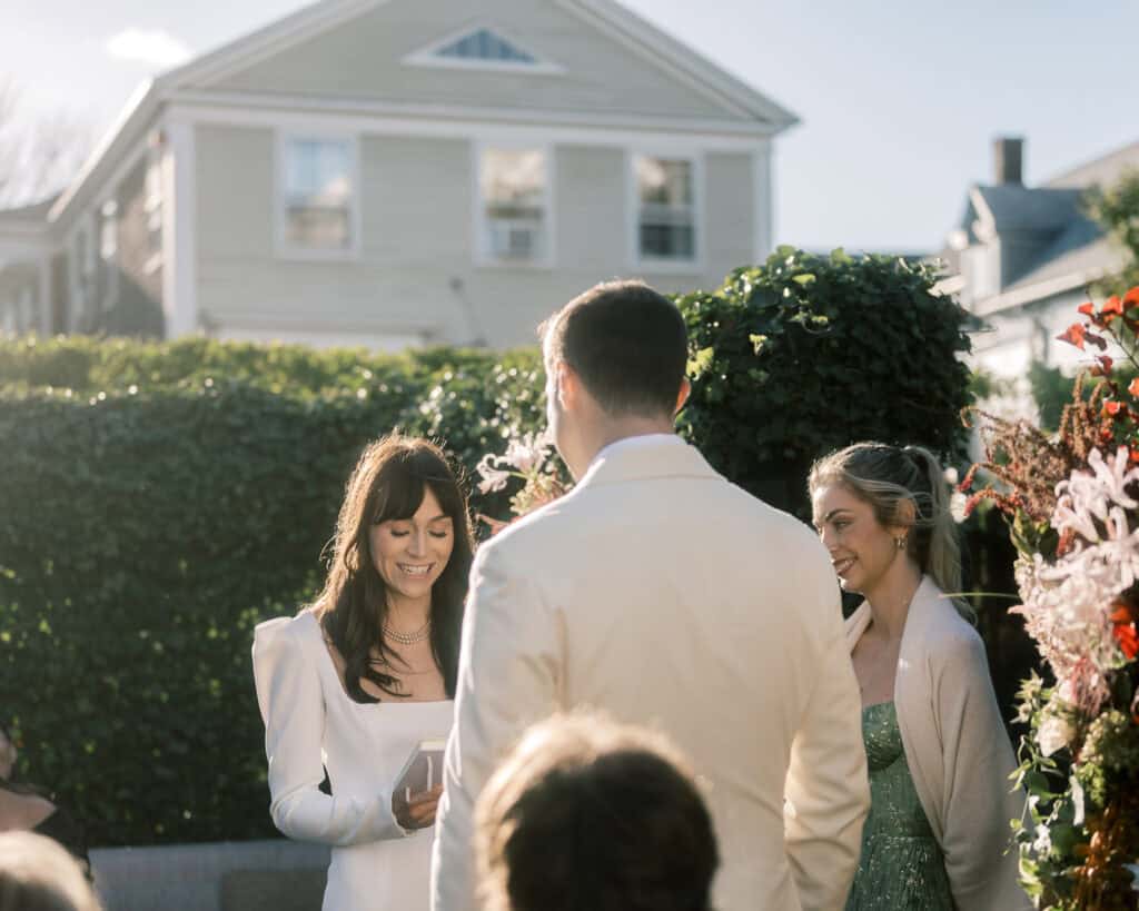 A bride in a white dress reads from a card to a groom in a light suit during an outdoor wedding ceremony, with another woman standing nearby—captured beautifully by Nantucket Wedding Photographer Kaity Farrell.