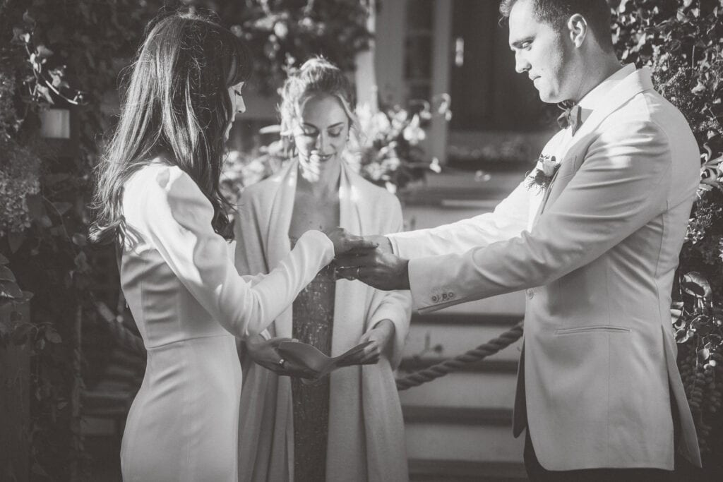 A couple exchanges rings during a wedding ceremony while an officiant stands between them, all dressed in formal attire, beautifully captured by Nantucket Wedding Photographer Kaity Farrell.