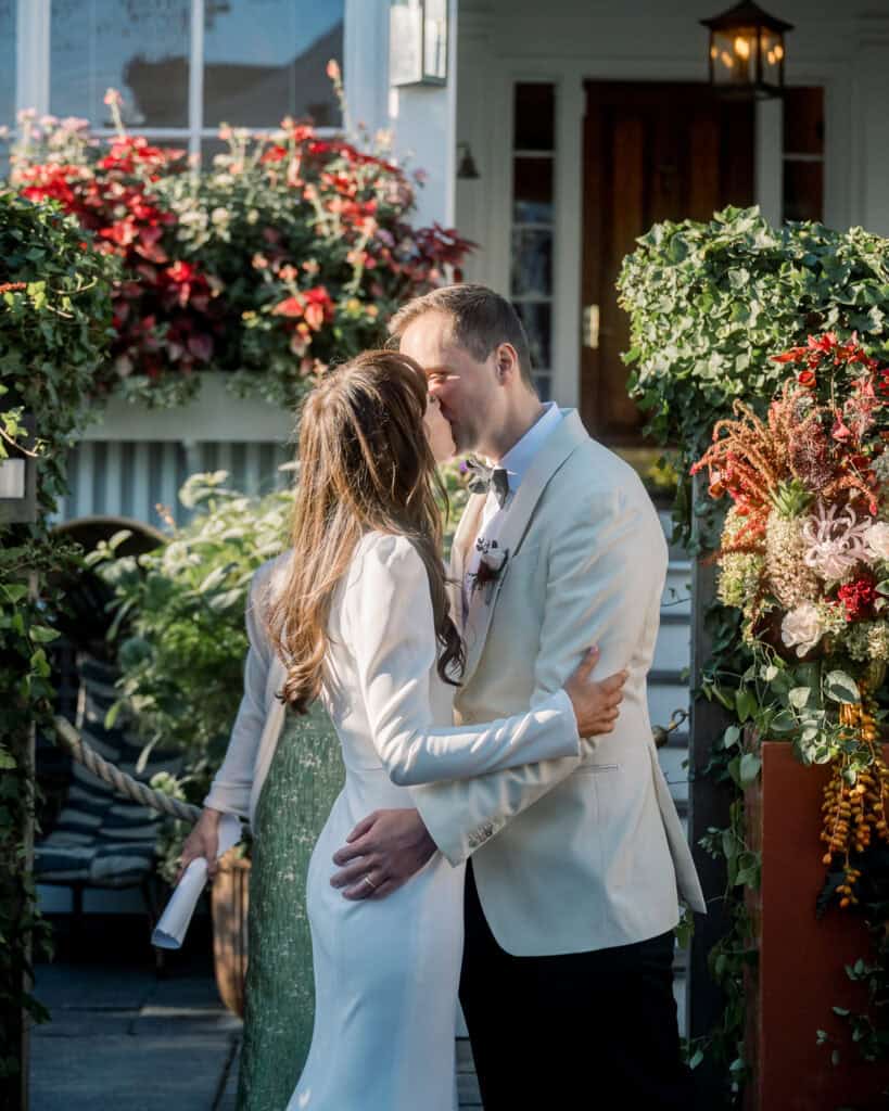 A couple in formal attire shares a kiss outdoors, surrounded by greenery and flowers, with a house entrance in the background—captured beautifully by Nantucket wedding photographer Kaity Farrell.