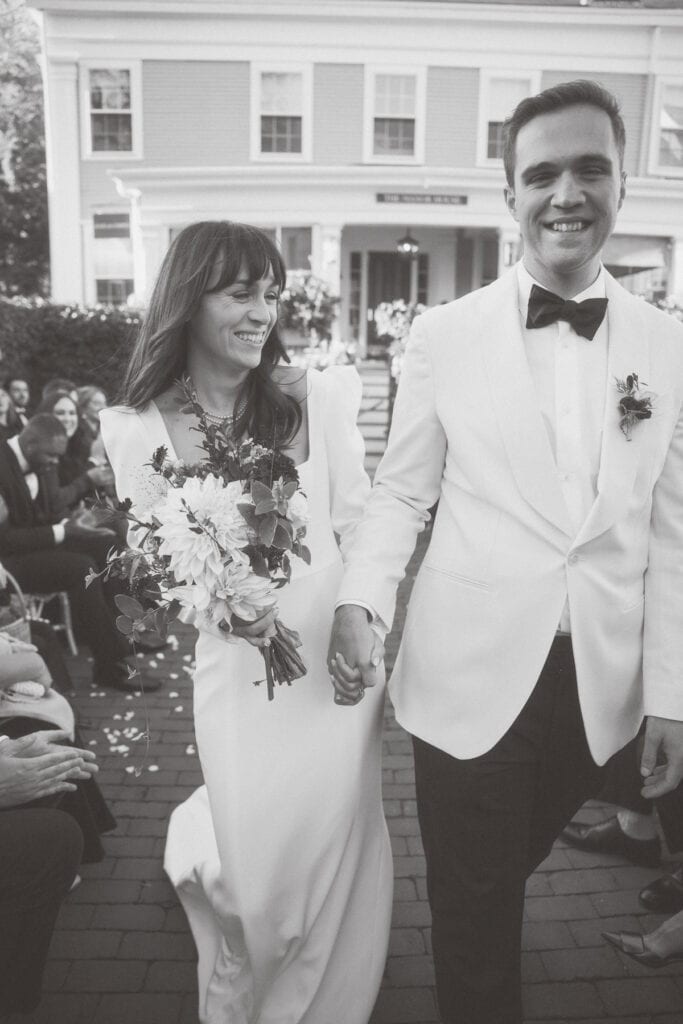 A bride and groom, both in white attire, walk hand-in-hand down an outdoor aisle, smiling, with guests seated on either side.