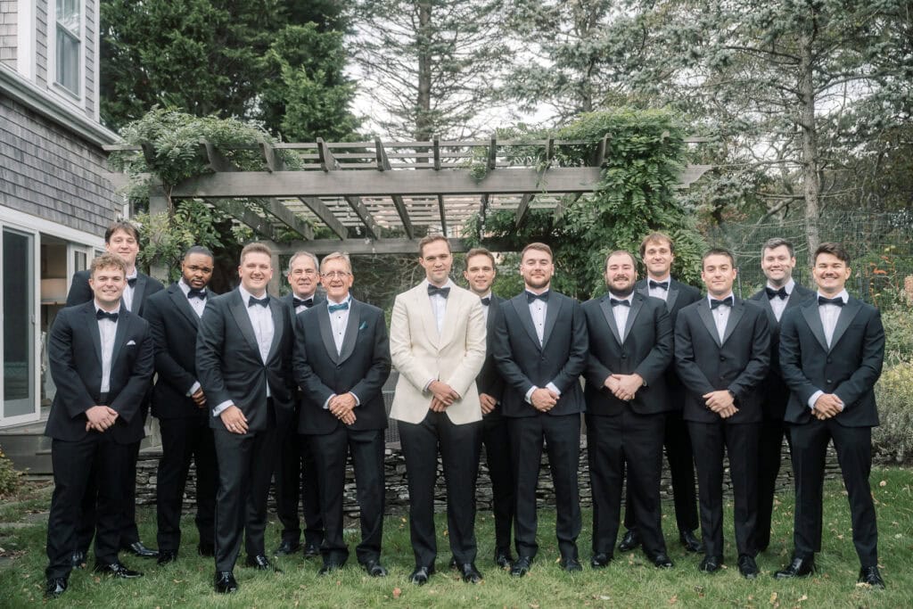 A Nantucket wedding party of thirteen men in tuxedos pose for a formal outdoor photo; the groom in the center wears a white jacket, while the others wear black, beautifully captured by Nantucket Wedding Photographer Kaity Farrell.