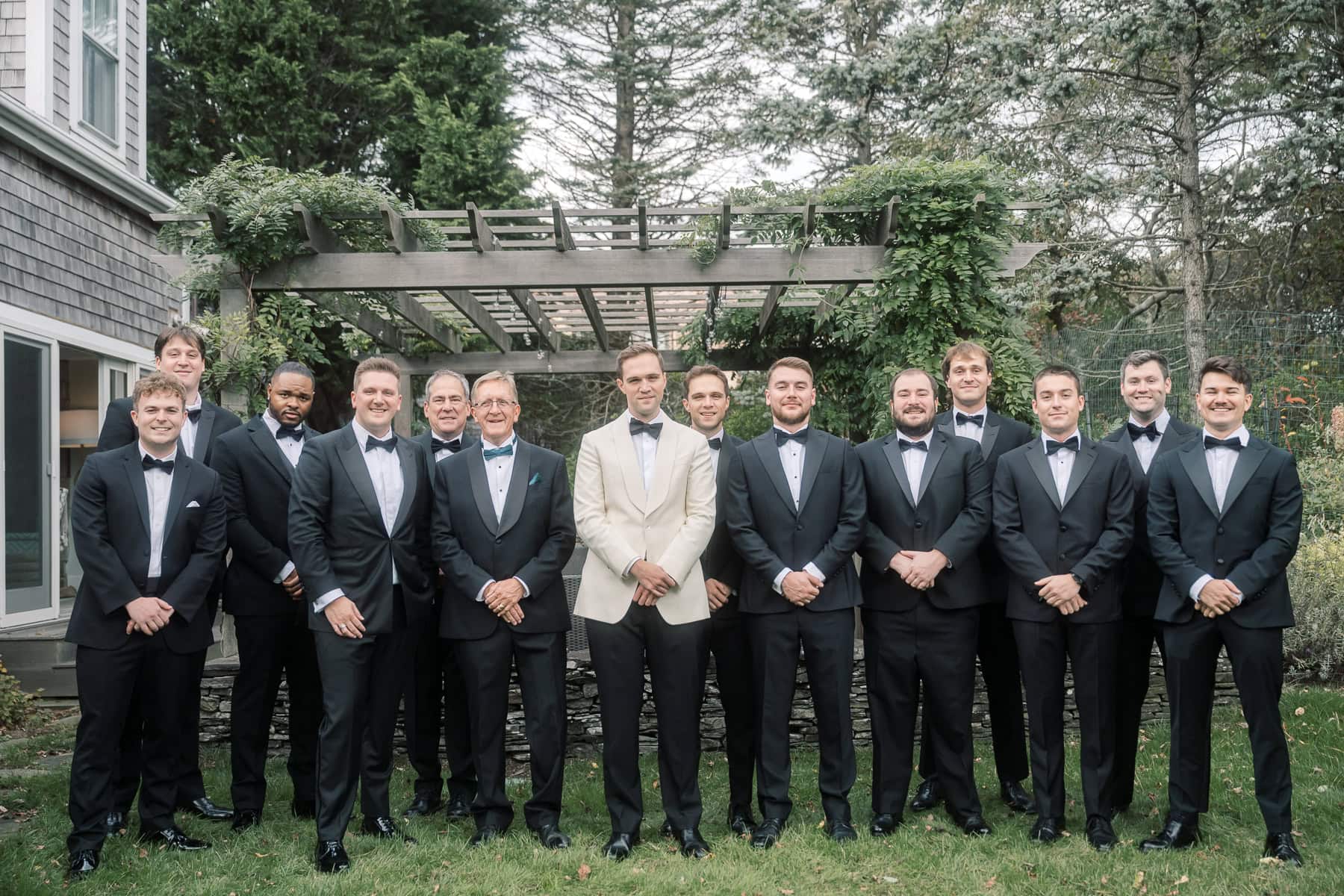 A Nantucket wedding party of thirteen men in tuxedos pose for a formal outdoor photo; the groom in the center wears a white jacket, while the others wear black, beautifully captured by Nantucket Wedding Photographer Kaity Farrell.