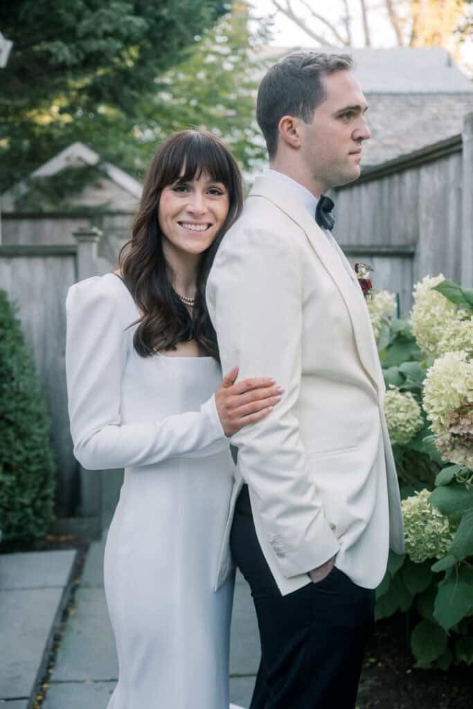 The bride in a white dress smiles, her hand on the arm of the groom in a white tuxedo jacket, as they stand near greenery and a wooden fence. Captured by Nantucket Wedding Photographer Kaity Farrell.