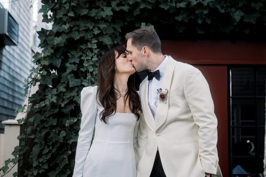 A bride and groom in formal attire share a kiss outdoors in front of a leafy wall and a red building, captured beautifully by Nantucket Wedding Photographer Kaity Farrell.