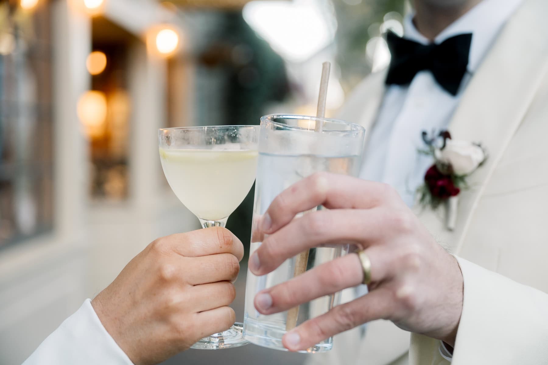 A bride and groom clink a cocktail glass and a wine glass in formal attire—one with a boutonniere and wedding ring—captured by Nantucket Wedding Photographer Kaity Farrell.