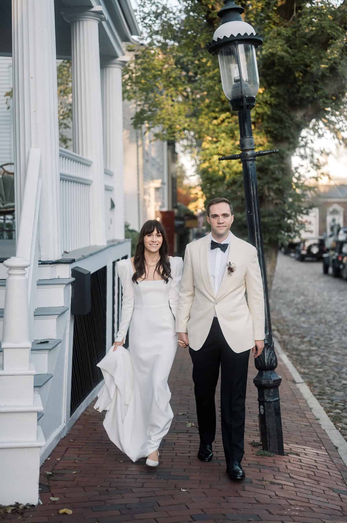 A bride in a white dress and a groom in a white tuxedo jacket walk hand in hand on a brick sidewalk beside a streetlamp and a house with columns.