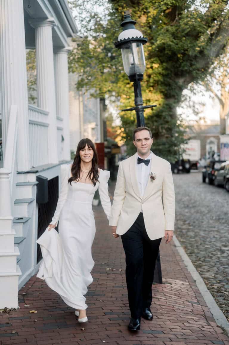 A bride and groom, both in white attire, walk hand in hand on a brick sidewalk beside a streetlamp in a residential neighborhood—captured beautifully by Nantucket Wedding Photographer Kaity Farrell.