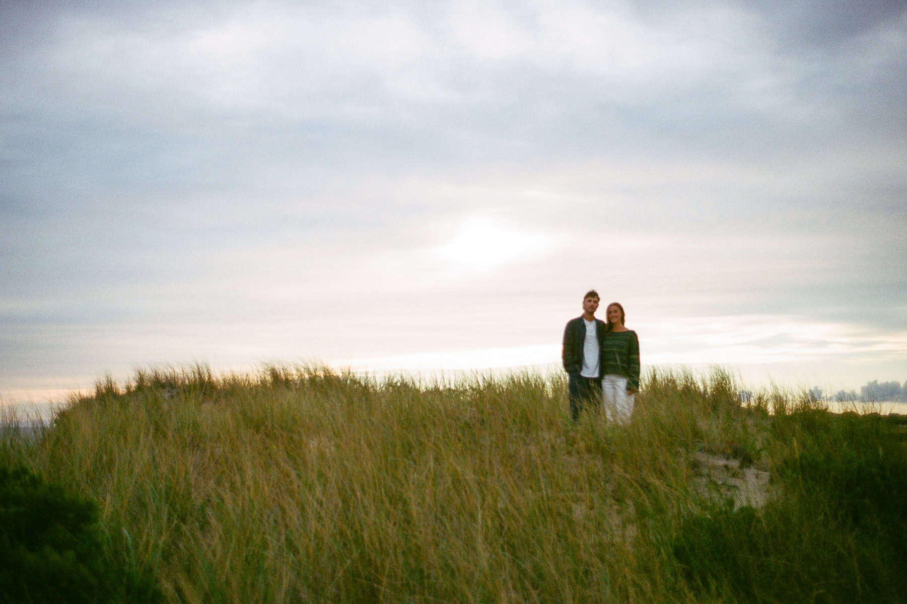 Two people stand together on a grassy sand dune under a cloudy sunset sky, captured during an engagement photo session by Nantucket wedding photographer Kaity Farrell.