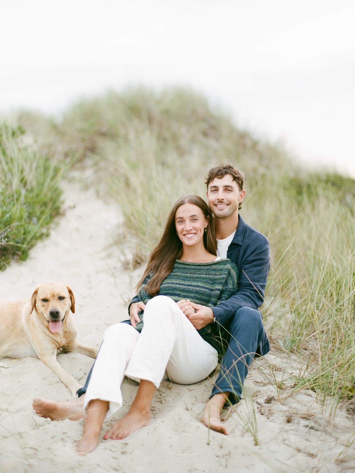 A couple sits together on sandy ground among grassy dunes, smiling at the camera, with a yellow labrador lying beside them—captured beautifully by Nantucket engagement photographer Kaity Farrell.