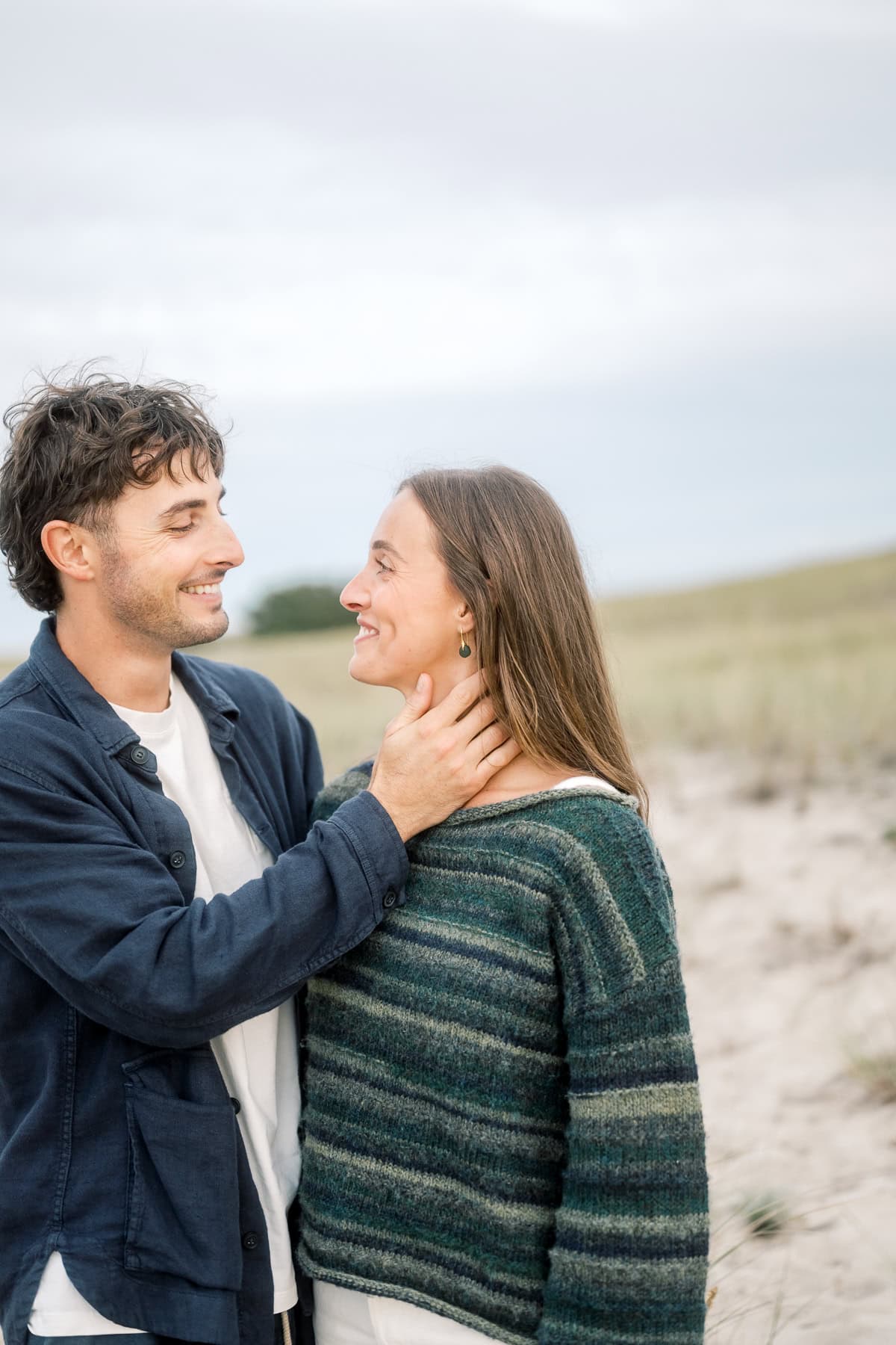 A man and woman stand close together outdoors on a sandy path, smiling at each other. Nantucket couples photographer Kaity Farrell captures the moment as the man gently holds the woman's neck, with grassy dunes and a cloudy sky in the background.