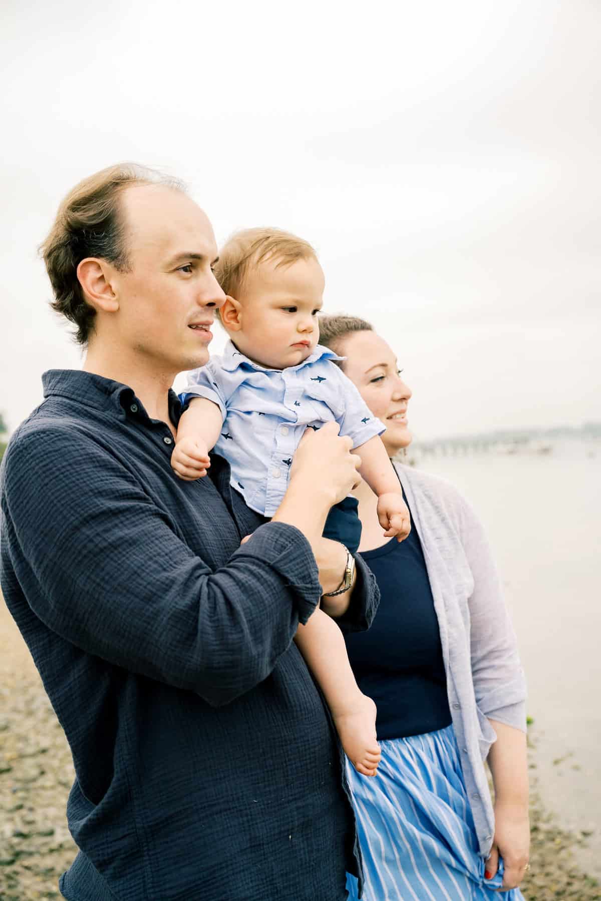 A man holds a baby while standing next to a woman on a rocky beach, all gazing into the distance—a beautiful Nantucket family moment captured by photographer Kaity Farrell.