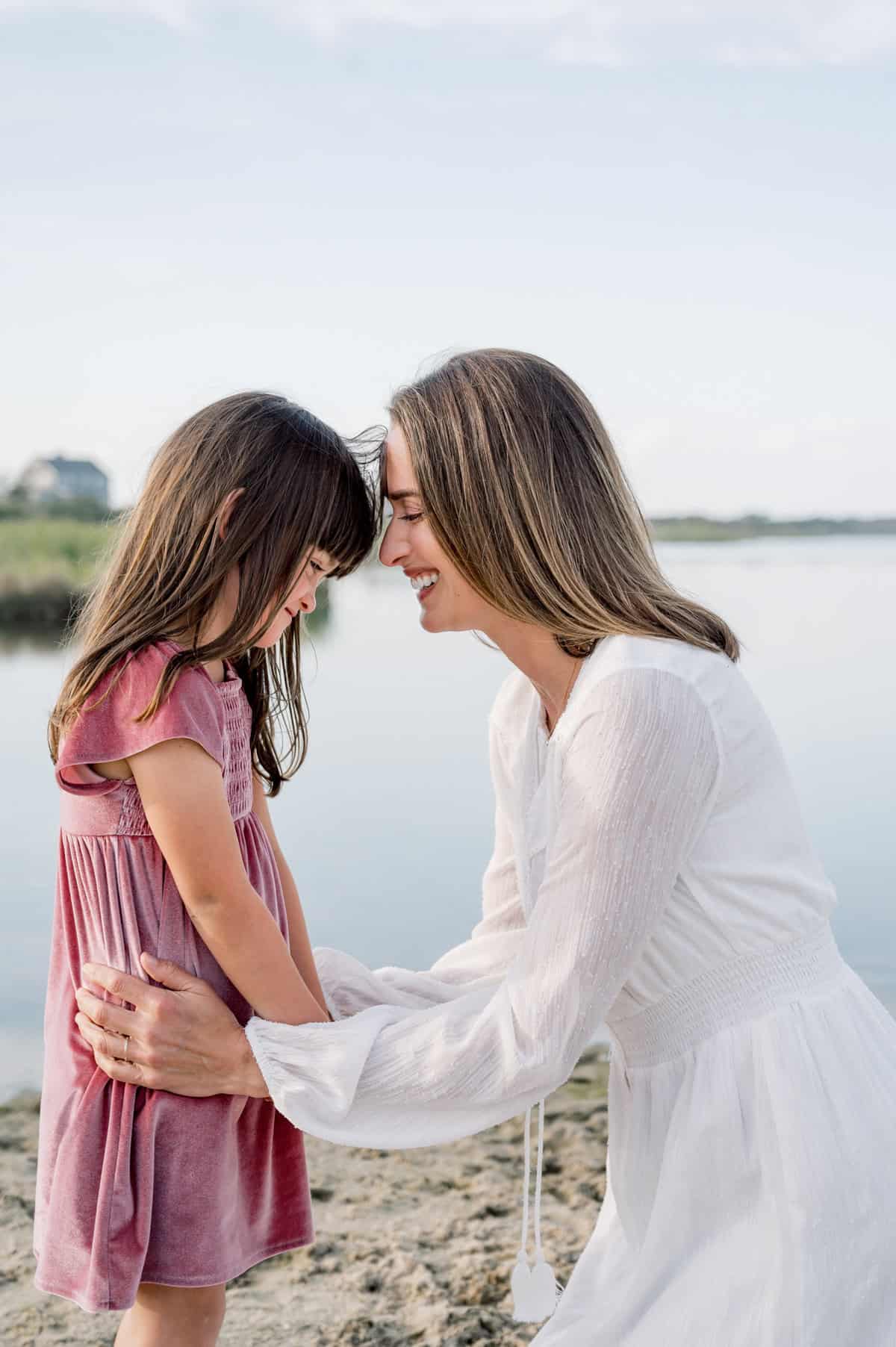A mother in a white dress kneels and smiles, gently holding the arms of her young daughter in a pink dress by a calm body of water—captured beautifully by Nantucket family photographer Kaity Farrell.