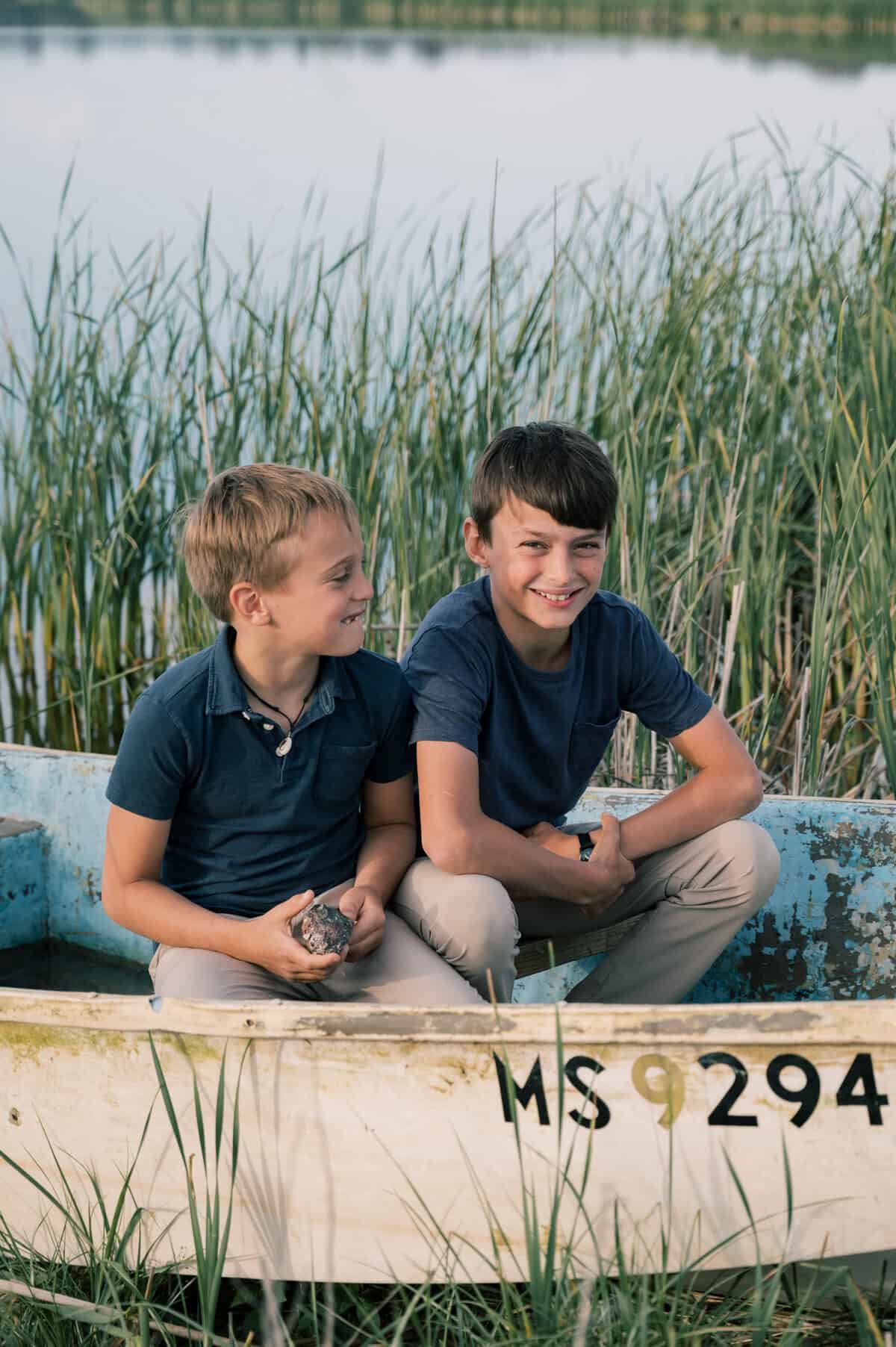 Two boys are sitting in an old white rowboat near tall grass by the water, both smiling and wearing blue shirts and light pants—captured beautifully by Nantucket family photographer Kaity Farrell.