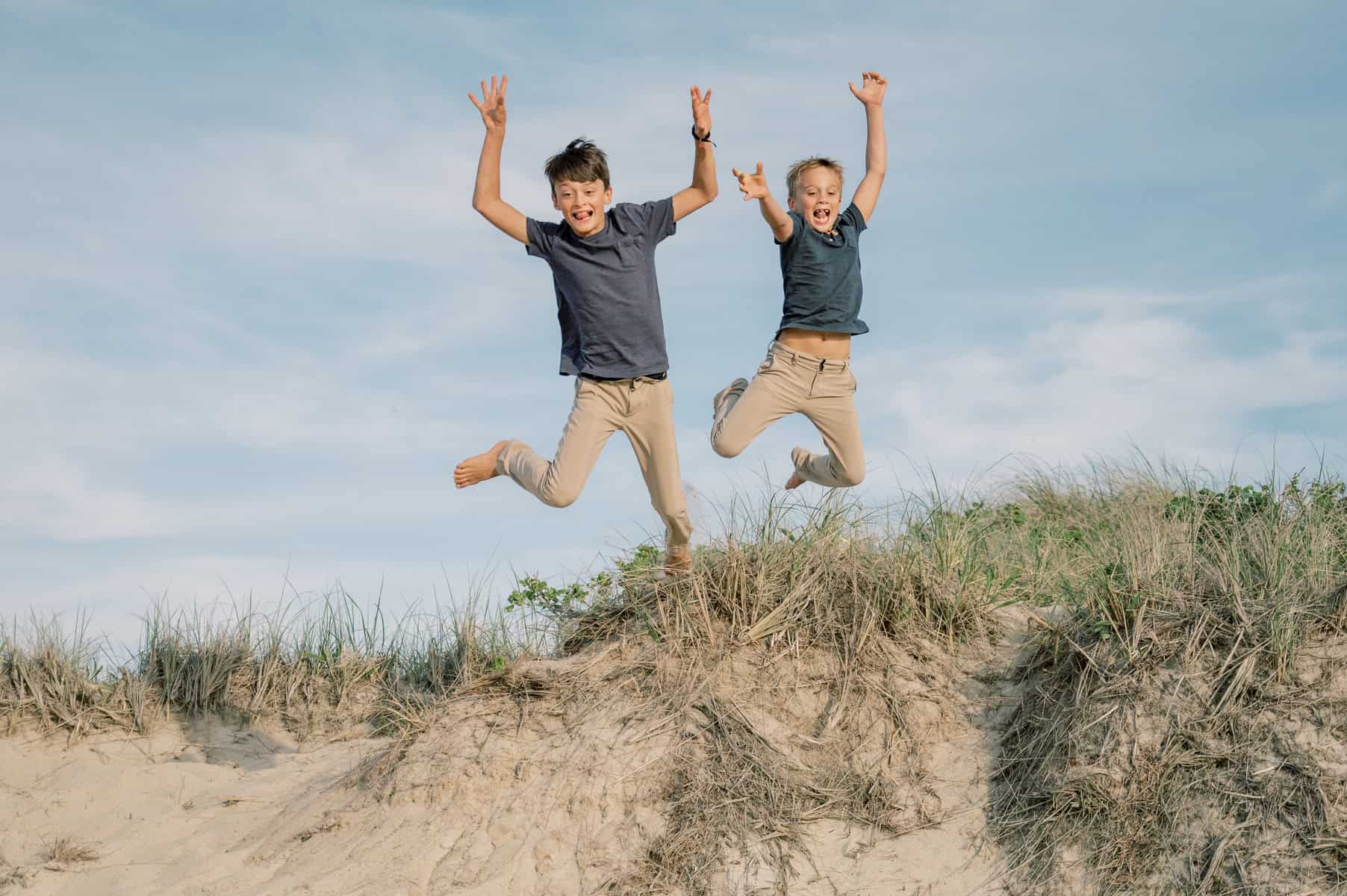 Two children in blue shirts and beige pants leap with arms raised above a sandy dune under a blue sky—captured by Nantucket photographer Kaity Farrell during a joyful Nantucket family photo session.