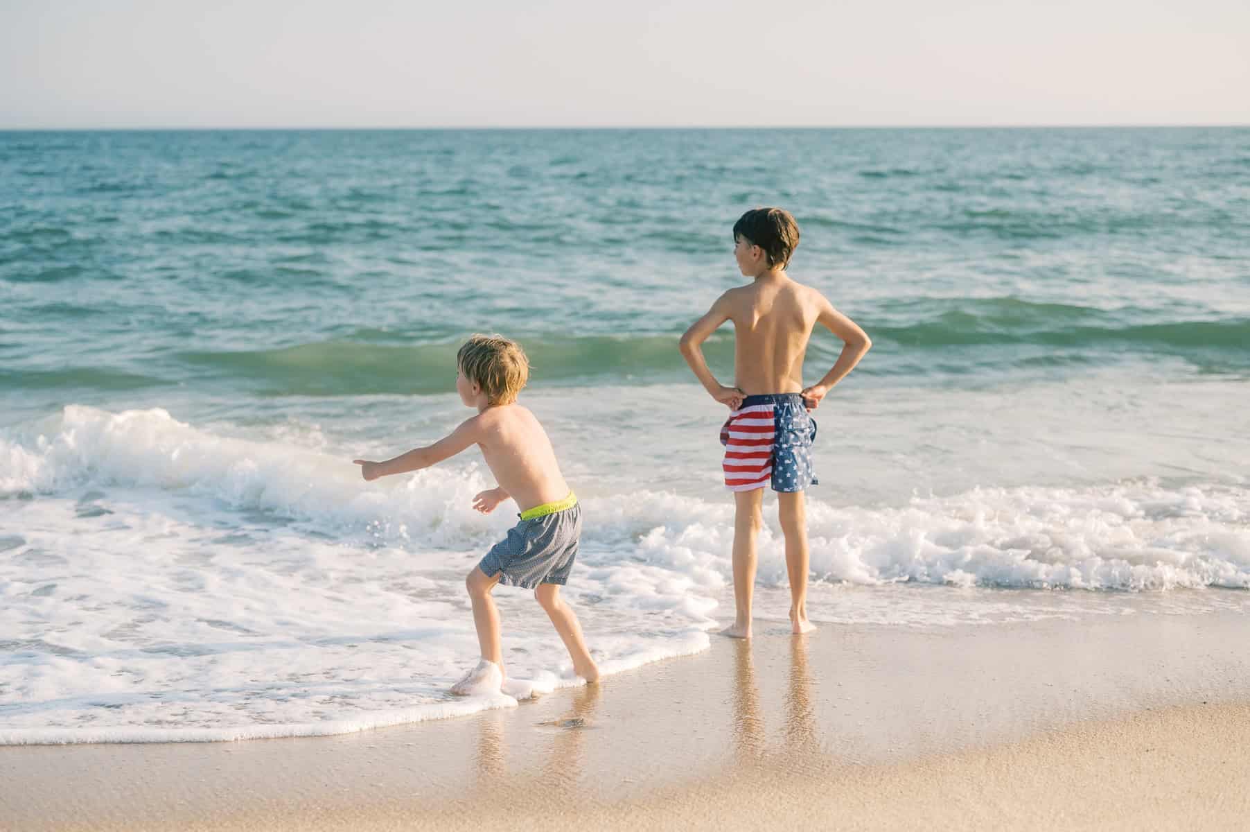 Two boys in swim trunks stand at the edge of the ocean at Miacomet Beach in Nantucket; one tosses something into the water while the other looks on during a Nantucket family photo session with photographer Kaity Farrell.